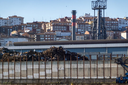 View of a large scrap metal yard with industrial chimneys and residential apartment buildings on a hillside under a clear blue sky in Greater Bilbao, Basque Country, Spain.