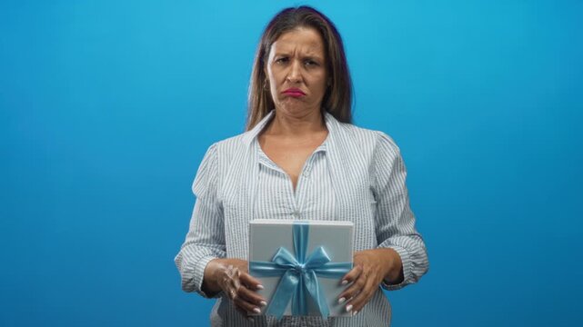 Woman holding a wrapped gift with blue ribbon in studio, hands visible and frowning; quiet disappointment.