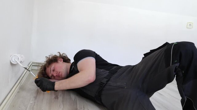 Exhausted renovation worker keeps painting wall while lying on floor showing burnout fatigue and nonstop apartment repair during home improvement process