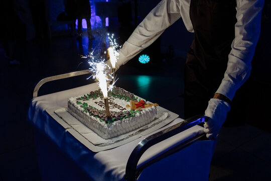 Waiter serving a beautiful festive graduation cake with bright burning fireworks in a dark banquet hall during a celebration.