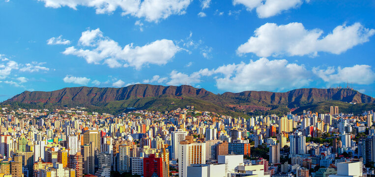Panoramic view of Belo Horizonte skyline with the iconic Serra do Curral mountains under a blue cloudy sky. Urban landscape featuring dense high-rise buildings and natural mountain preservation.