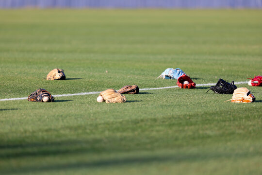 Multiple baseball gloves and baseballs rest on the grass beside a painted field line during pregame warmups at a high school baseball game in Olive Branch, Mississippi.