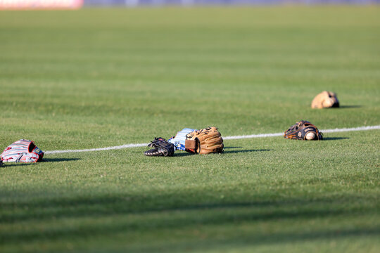 Multiple baseball gloves and baseballs rest on the grass beside a painted field line during pregame warmups at a high school baseball game in Olive Branch, Mississippi.