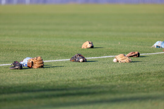 Multiple baseball gloves and baseballs rest on the grass beside a painted field line during pregame warmups at a high school baseball game in Olive Branch, Mississippi.