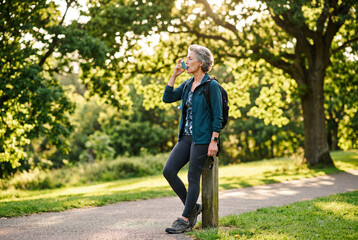 Obraz premium Mature woman with backpack standing outdoors using an asthma inhaler in a sunny green park