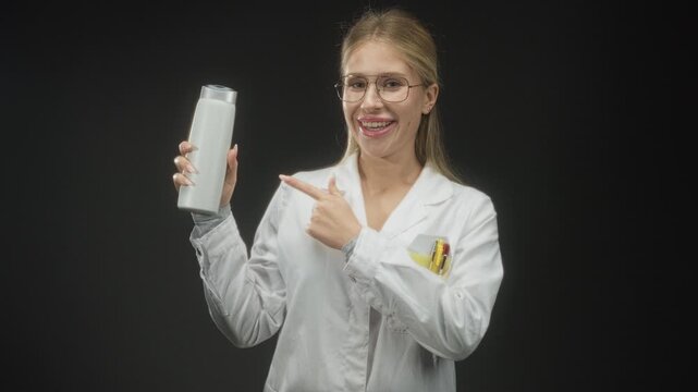 Woman in white coat pointing hand to white bottle while smiling in studio with dark backdrop; confidence product endorsement.