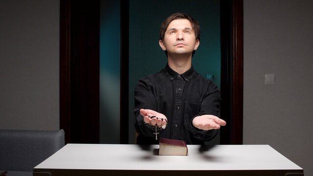 Young man sitting at a table looking up in prayer, holding a rosary in one hand and the other hand open in a quiet, intimate moment of devotion and hope toward god