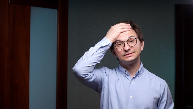 Man in a blue shirt and glasses looking stressed and disappointed, holding his hand to his forehead, feeling the effects of a headache, regret, or exhaustion