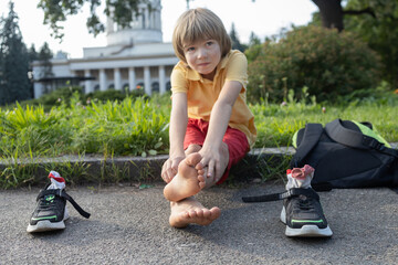 boy sits tiredly on the sidewalk, having removed his sneakers and backpack. Focus on his feet. The child is barefoot and sockless, trying to cool his feet in the hot weather and rest © Anna