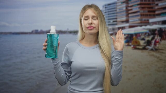 Woman holding mouthwash bottle, hand raised near nose and lips in a disgusted grimace by a seaside building with umbrellas and sand; disgust caution.