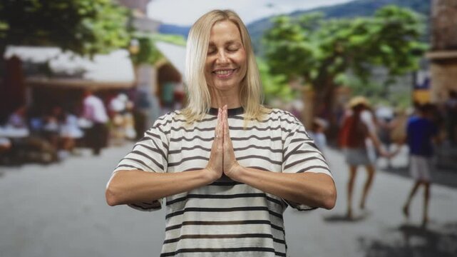 Blonde middle aged woman in striped shirt pressing hands together in namaste prayer gesture on street market; calm reflection.
