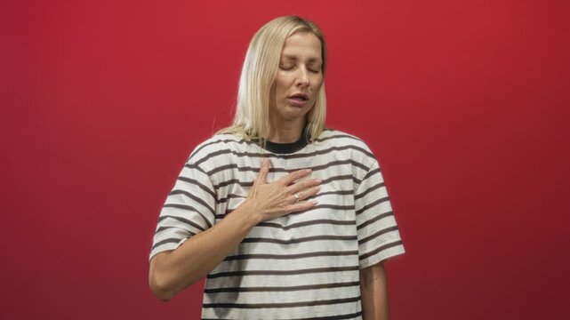 Woman in striped tshirt, blonde middleaged, gasping with hand on chest in studio against red backdrop; breath relief.