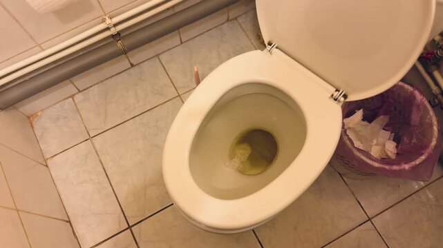 Dirty public toilet bowl showing urine, open lid and porcelain bowl stained yellow with crumpled tissue inside, tiled floor