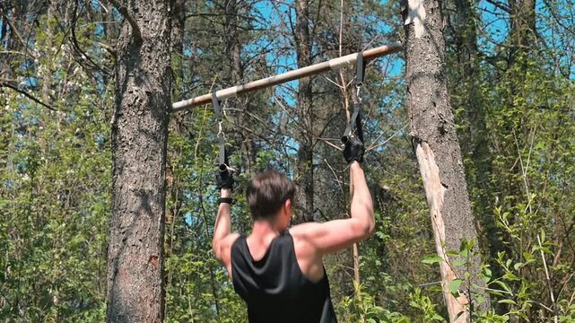 A man engages in pull-up exercises on a wooden bar hung between two trees in a forested area. He is wearing a sleeveless shirt and gloves, focusing on physical fitness and strength training in nature