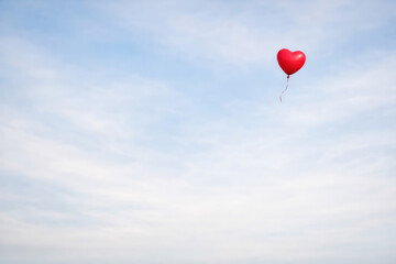 Fototapeta premium A single small red heart shaped balloon floating in a bright blue sky with light wispy clouds, representing love, joy, and simplicity