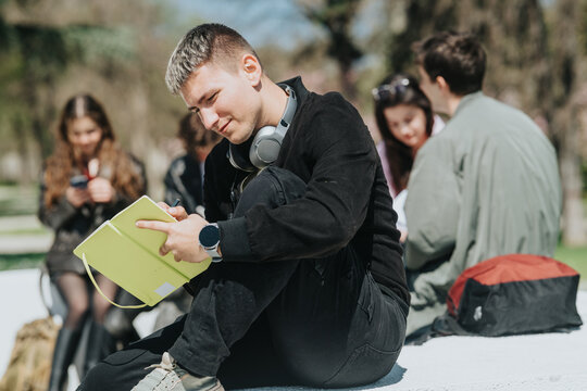 Young male student writing notes in a notebook while sitting outdoors with school friends nearby. A small group of students congregate on campus grounds as he studies and takes notes.