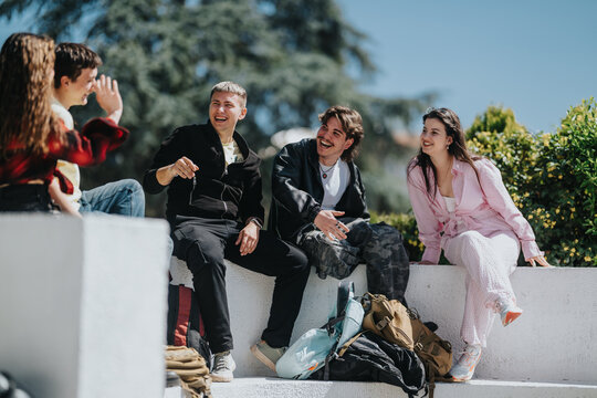 A group of school friends sit on a low outdoor wall smiling and talking together. Teen students socialize and laugh during a sunny break with backpacks nearby.