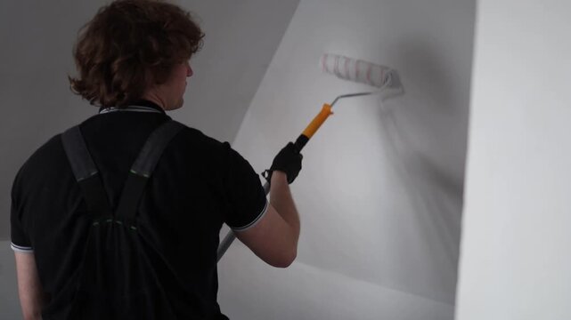 Young man repainting attic apartment walls from behind during costly home renovation project trying to refresh living space with fresh white paint