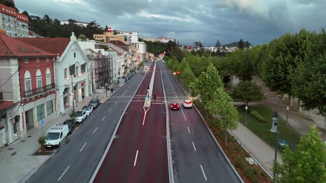 Drone shot looking down a wide modern boulevard with red bus lanes and vehicles alongside a scaffolded building under renovation with the Coimbra hillside in the background
