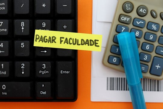 Post-it note with the words "Pagar Faculdade" (Paying College, in Portuguese) on a study desk with a calculator, computer, barcodes and calendar. Concept of education costs in Brazil.