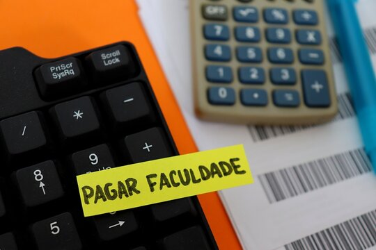 Post-it note with the words "Pagar Faculdade" (Paying College, in Portuguese) on a study desk with a calculator, computer, barcodes and calendar. Concept of education costs in Brazil.