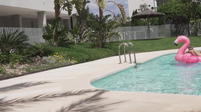 Little girl floating on a pink inflatable flamingo in a sunny outdoor swimming pool surrounded by tropical plants. sstkSummer lifestyle video showing family vacation, poolside leisure, summer fun and