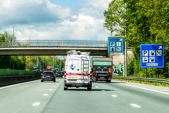Wetteren, Belgium - April 20, 2026: Traffic on the E40 highway near Wetteren, showing service area road signs for parking, fuel, and restaurants under a bridge with lush green trees on the side.