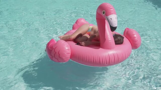 Little girl floating on a pink flamingo inflatable ring in a swimming pool on a sunny summer day. Fun childhood leisure, vacation lifestyle, and relaxing pool activity concept