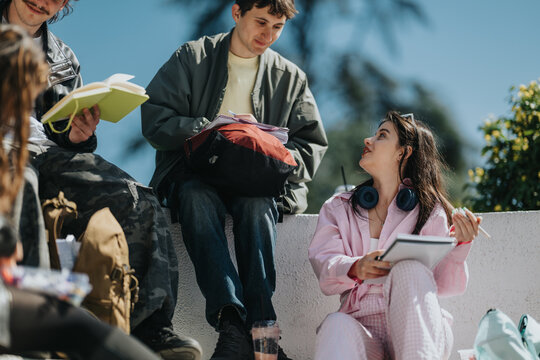 High school students sit together outdoors studying and talking with notebooks and headphones. A casual group of friends share notes and ideas while relaxing on a campus wall.