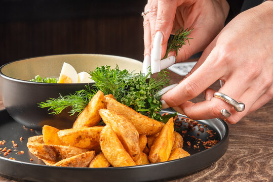 A pile of crispy potato wedges served on a plate with fresh dill and a side dish, with hands decorating it.