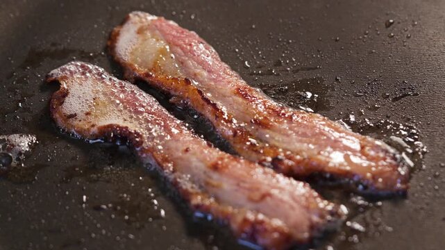 Two bacon strips sizzling on a hot griddle with grease splatter golden brown crispy breakfast pork closeup. 