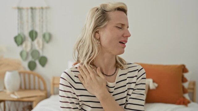 Woman rubbing neck and shoulder on bed in studio bedroom set, wearing striped shirt with pillows and macrame wall hanging visible; discomfort healing.