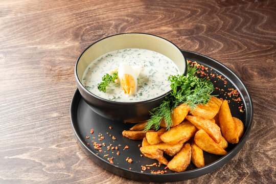 Okroshka cold soup in a black bowl with boiled egg and fried potato wedges on a dark plate, wooden background.