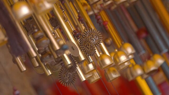 A multitude of shiny gilded tubular wind chimes with bells and engraved Hindu Om signs hang in a row and sway in the wind, producing melodic sounds
