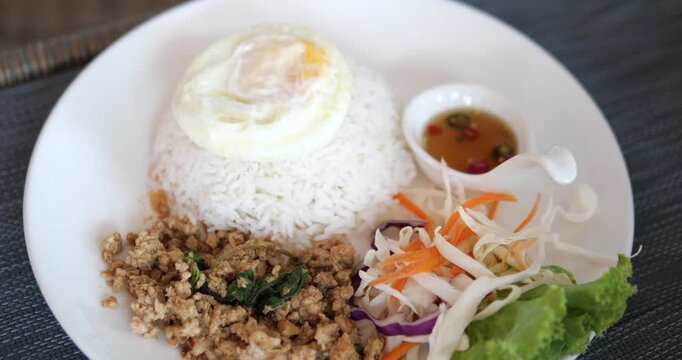 Cinematic close-up of traditional Thai stir-fried minced pork with holy basil (Pad Kra Prow) served with steamed rice, fried egg, and fresh salad on a white plate