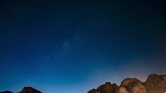 Night timelapse of Table Mountain and Milky Way near Cape Town A bright moon rises, city lights glow, and a small lit structure sits atop a rugged peak.
