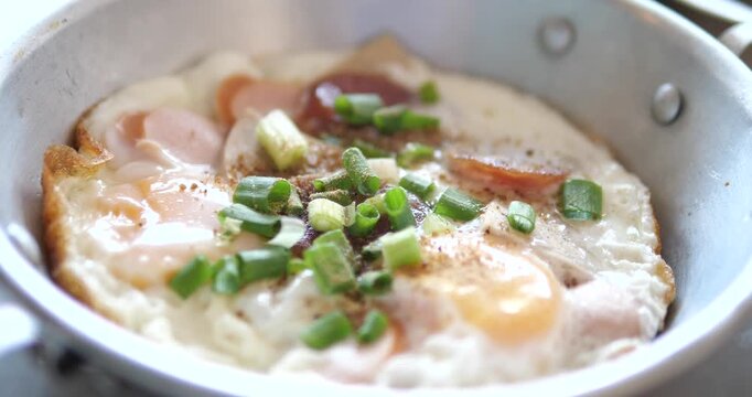 Cinematic close-up of traditional Asian pan-fried eggs (Kai Kata) topped with Chinese sausage, Vietnamese pork roll, and chopped spring onions in a small stainless steel pan, morning breakfast concept
