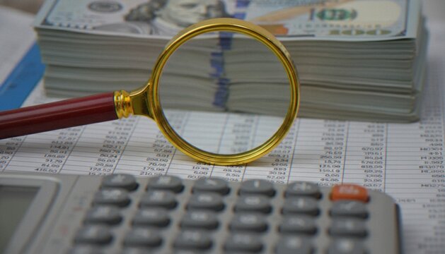 A close-up shot featuring a gold-rimmed magnifying glass resting on financial documents, with a stack of US hundred-dollar bills and a calculator in the blurred background.
