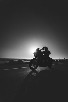 Motorcycle and Rider Silhouette Against Glowing Sunset on Coastal Highway in Black and White
