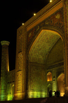 Sher-Dor Madrasah facade illuminated at night at Registan Square in Samarkand. Intricate tilework and massive iwan portal glow in golden and green light. Ideal for travel publications and cultural.