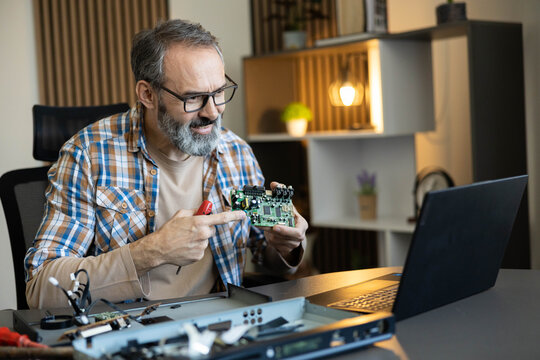 Man in checkered shirt holding a motherboard, explaining repair steps while consulting laptop during tech tutorial at desk