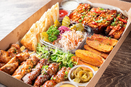A cardboard box filled with a variety of BBQ foods, including skewered meats, ribs, potato wedges, flatbread, pickles, coleslaw, and dipping sauces