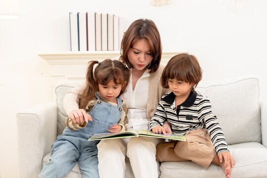Asian single mother guides daughter, son through page activity, explaining images step by step. Homeschooling learning game strengthens trust, patience, emotional support, family happiness indoors.