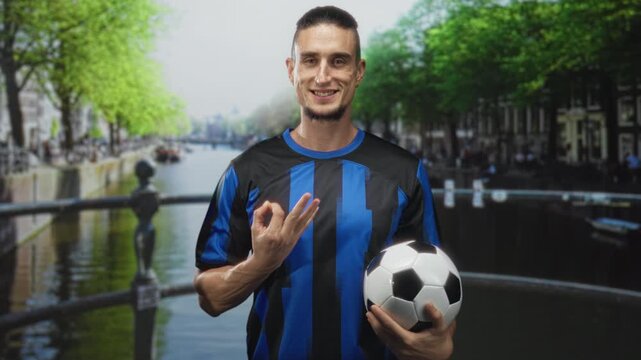 Man holding soccer ball and making ok sign on street by canal bridge, wearing blue striped jersey, smiling; confidence team spirit.