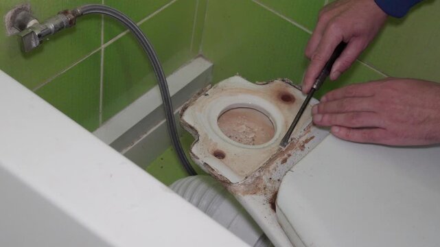 A plumber dismantles an old toilet in the bathroom. Close-up of the hands of a craftsman who uses a screwdriver or removes silicone from the dirty base of an old ceramic toilet. The process of plumbin