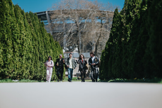 Five school friends walk together along a tree-lined campus path carrying backpacks and books. A casual student group strolls between tall hedges toward a building in the background.
