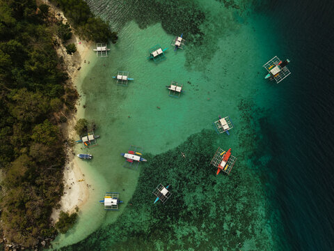 Aerial drone view of CYC Beach in Coron, Philippines, featuring crystal-clear turquoise waters, traditional Filipino outrigger boats and a tropical shoreline surrounded by lush island vegetation in Pa
