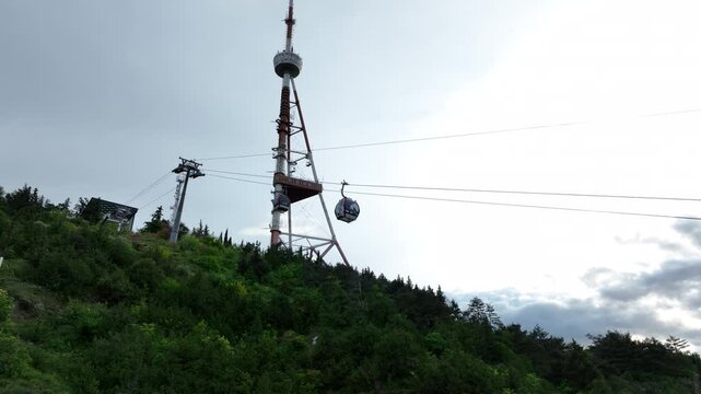 Aerial view of the modern cable car system in Tbilisi, Georgia, showing the upper station building, ropeway cables, and lush green hills under a dark cloudy sky
