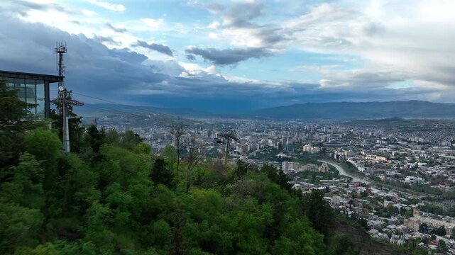 Aerial view of the modern cable car system in Tbilisi, Georgia, showing the upper station building, ropeway cables, and lush green hills under a dark cloudy sky