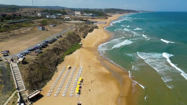 Beautiful drone tracking shot of a structured public beach with sun loungers and umbrellas bordered by a lush green hill landscape and clean emerald sea waves crashing on the sand